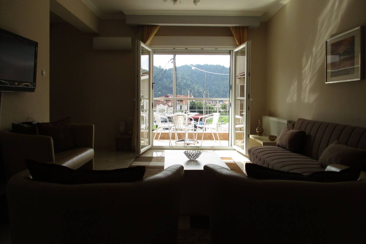 Een woonkamer met een bank, een televisie en uitzicht op een balkon met tuinmeubilair.