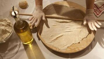 Une personne est en train d’étaler de la pâte sur une planche en bois à l’aide d’un rouleau à pâtisserie. Une bouteille d’huile et un bol de farine sont posés sur la table.