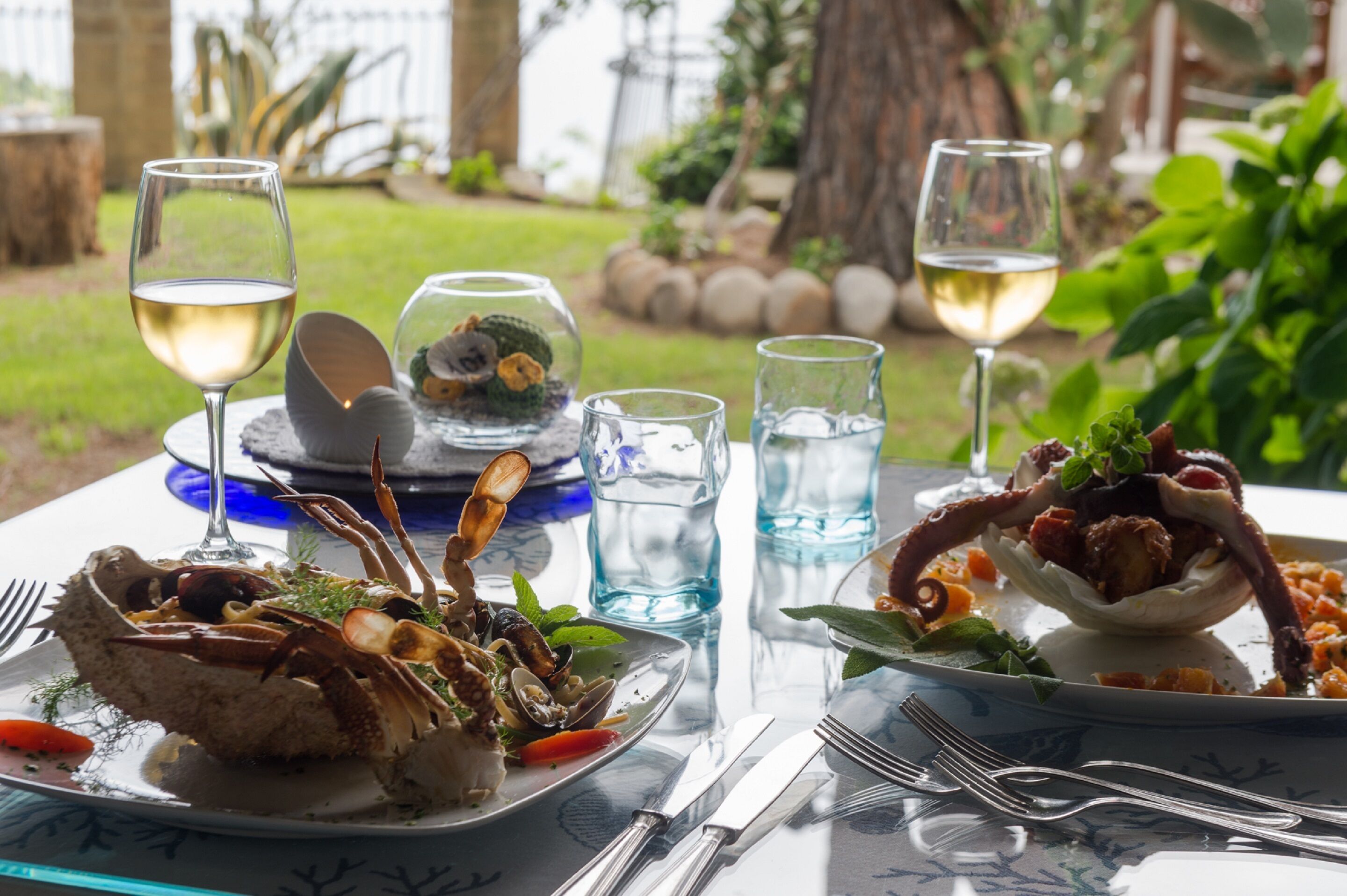 A table set with seafood dishes, wine glasses, and water glasses, with a view of a garden and a tree in the background.