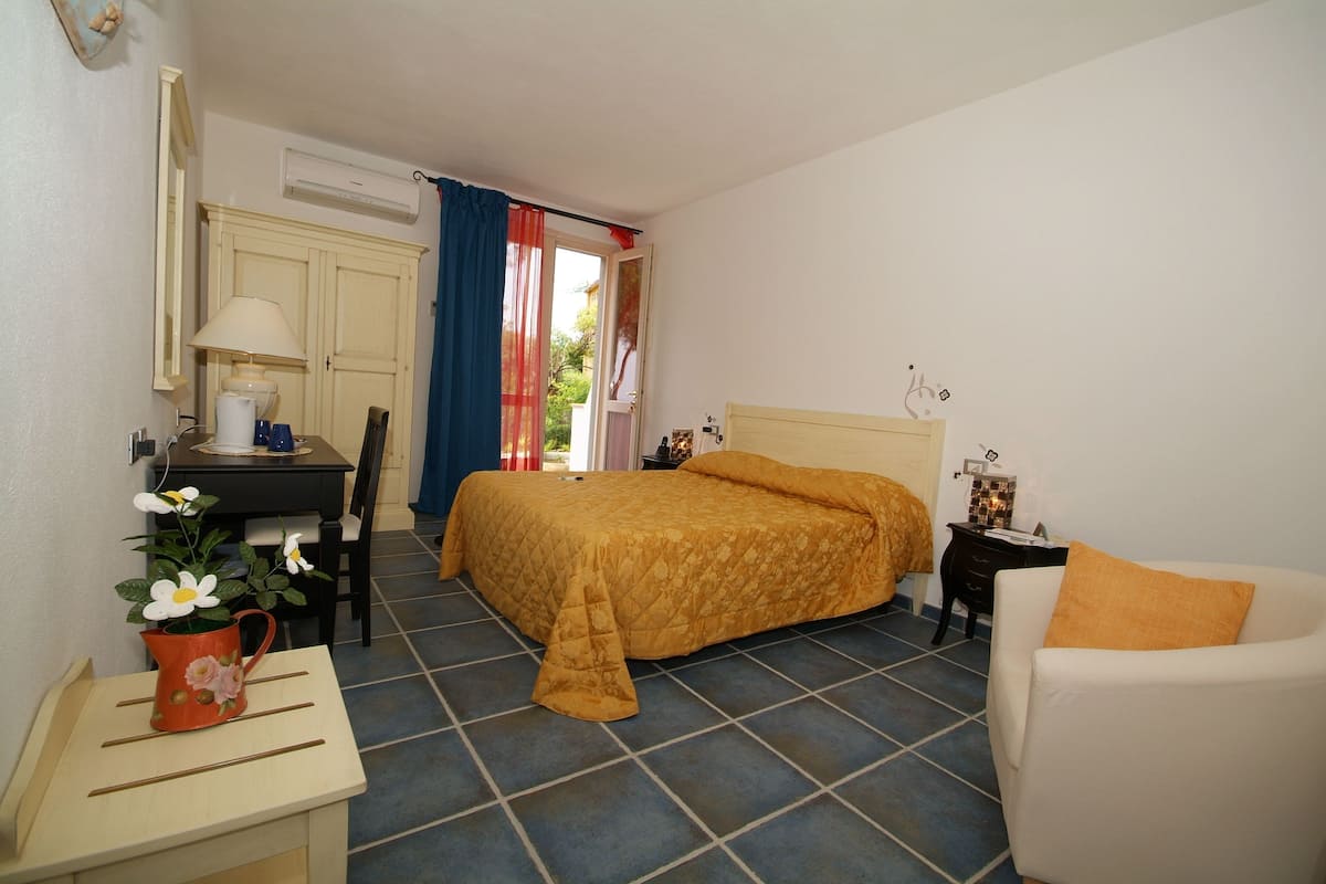 A bedroom with a bed, a desk, a chair, a bedside table, and a small table with a plant.