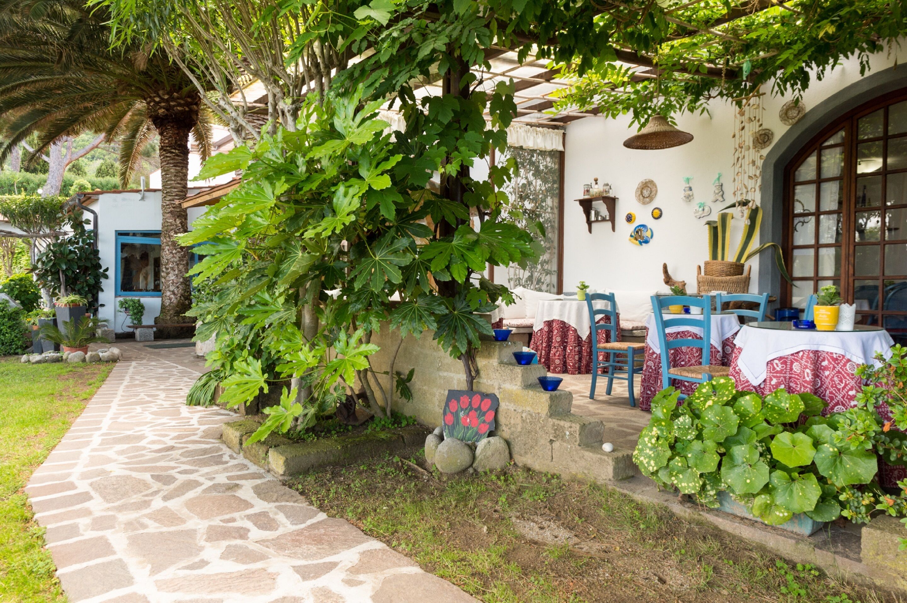 A stone pathway leading to a rustic outdoor seating area with a table and chairs, surrounded by lush greenery and a decorative wall.