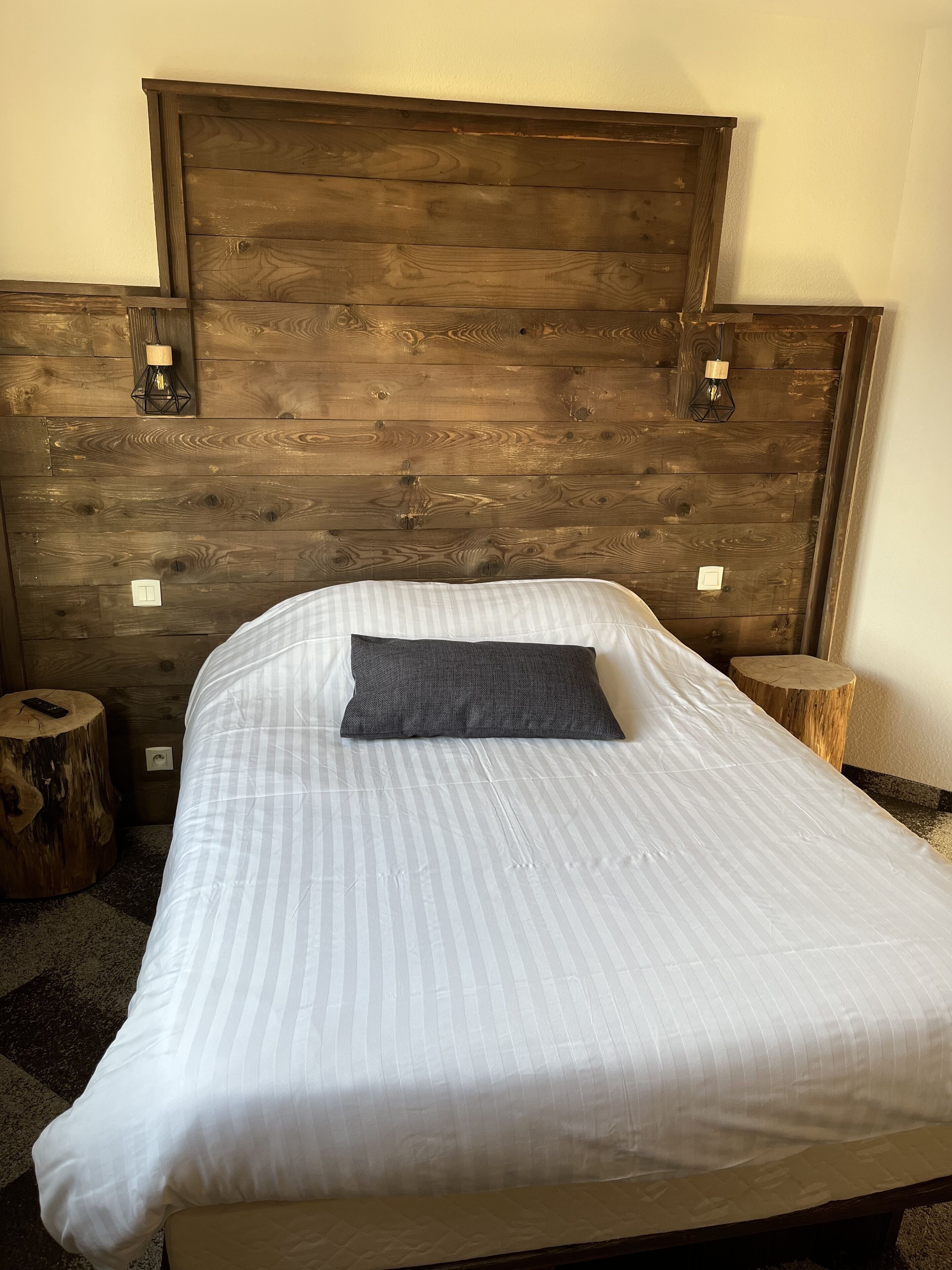 A bed with a wooden headboard, a striped white bedspread, and a dark pillow. There are two wooden nightstands with lamps and a small wooden stool.