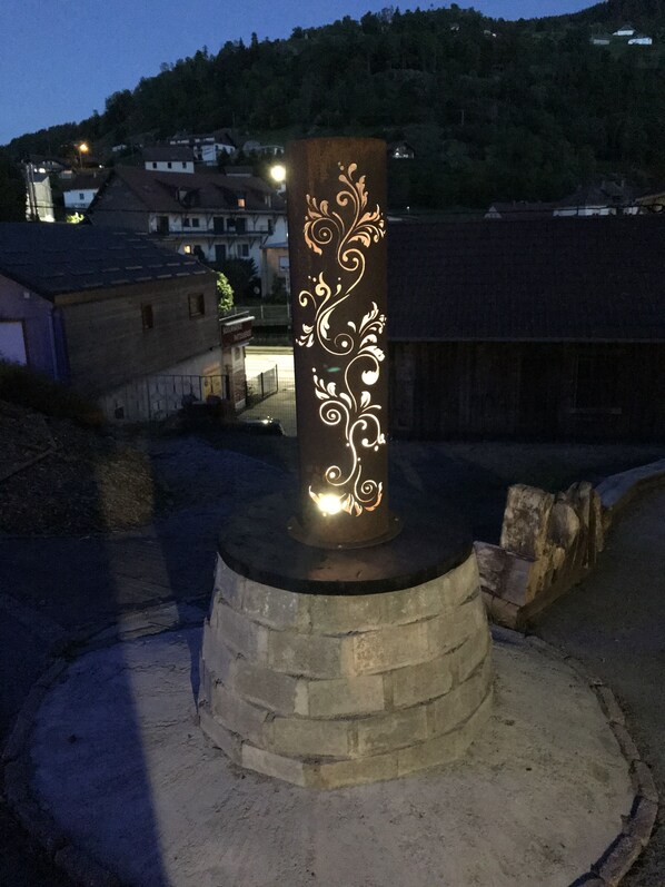 A cylindrical monument with intricate white designs, surrounded by a circular stone base, set against a backdrop of buildings and a hill.