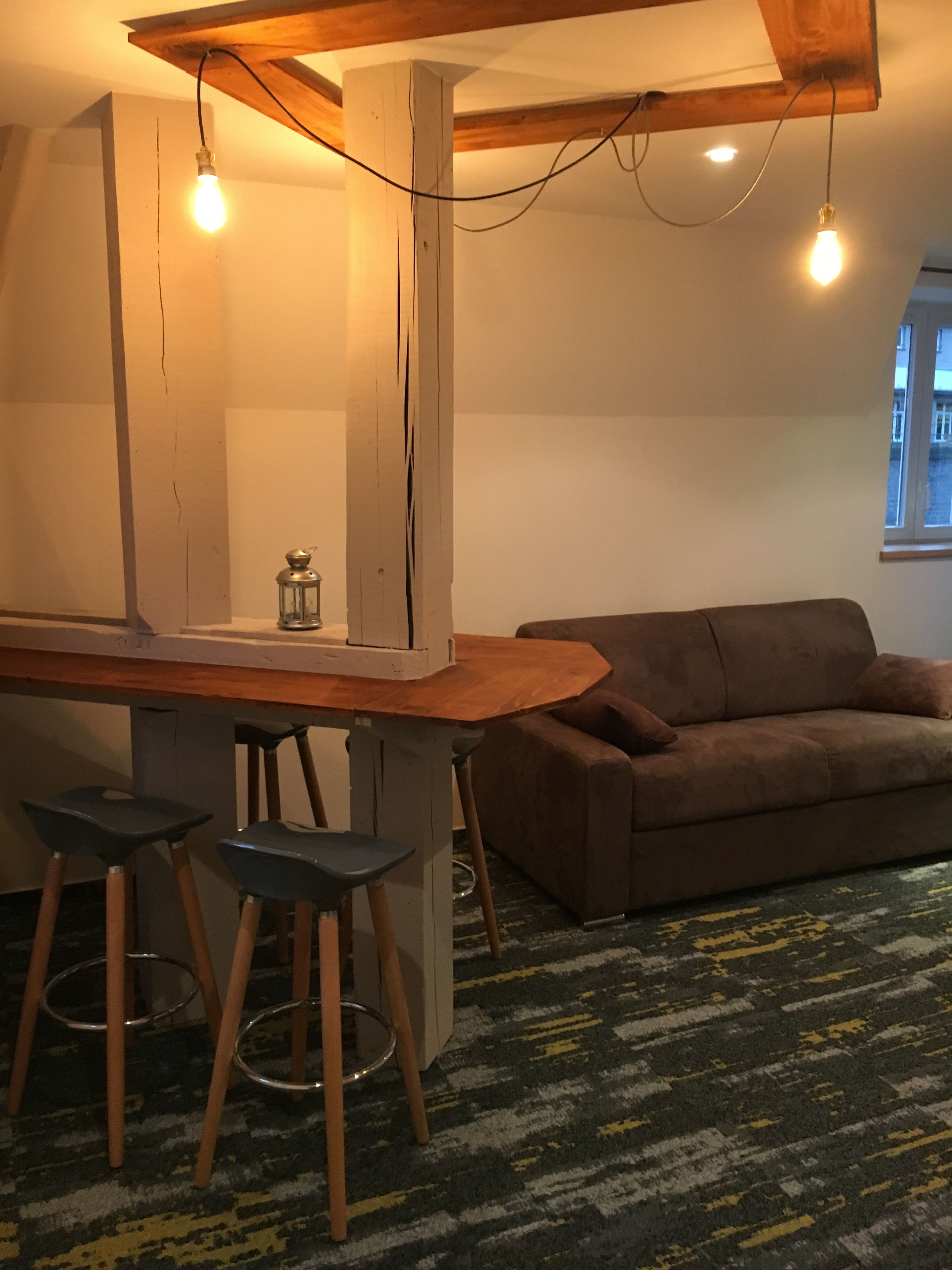 Ein gemütlicher Wohnbereich mit einer Holzbar, zwei Barhocker, einem braunen Sofa und einem gemusterten Teppich.