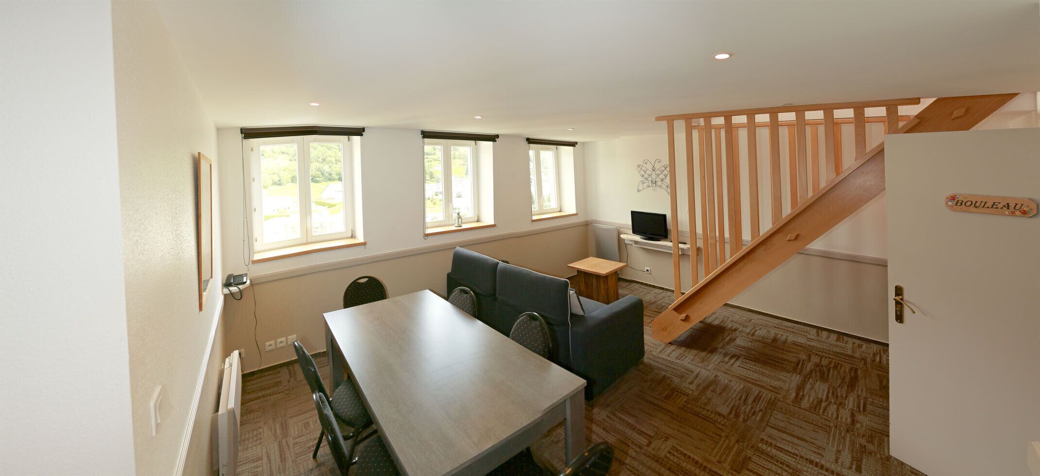 Una habitación con una mesa de comedor, dos sillas, un sofá, una mesita y un televisor. Hay tres ventanas y una escalera de madera.