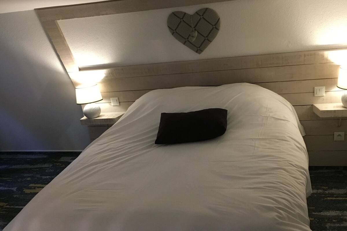 A hotel room with a bed, two bedside lamps, a heart-shaped wall decoration, and a nightstand.