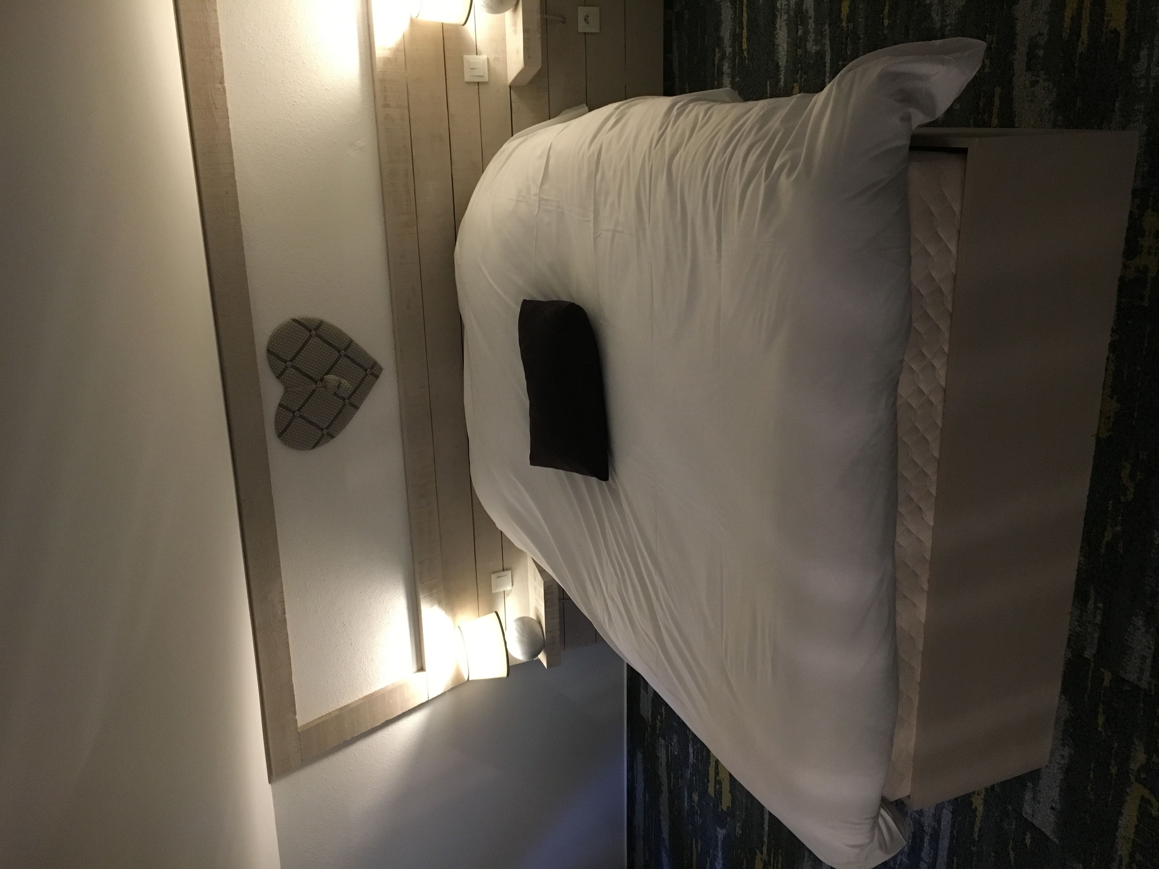 A bed with a white mattress and pillows, a small black object on the bed, and a wall-mounted light fixture.
