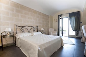 Quarto com cama, duas mesas de cabeceira com abajures, uma cadeira e vista para o exterior através de uma porta.