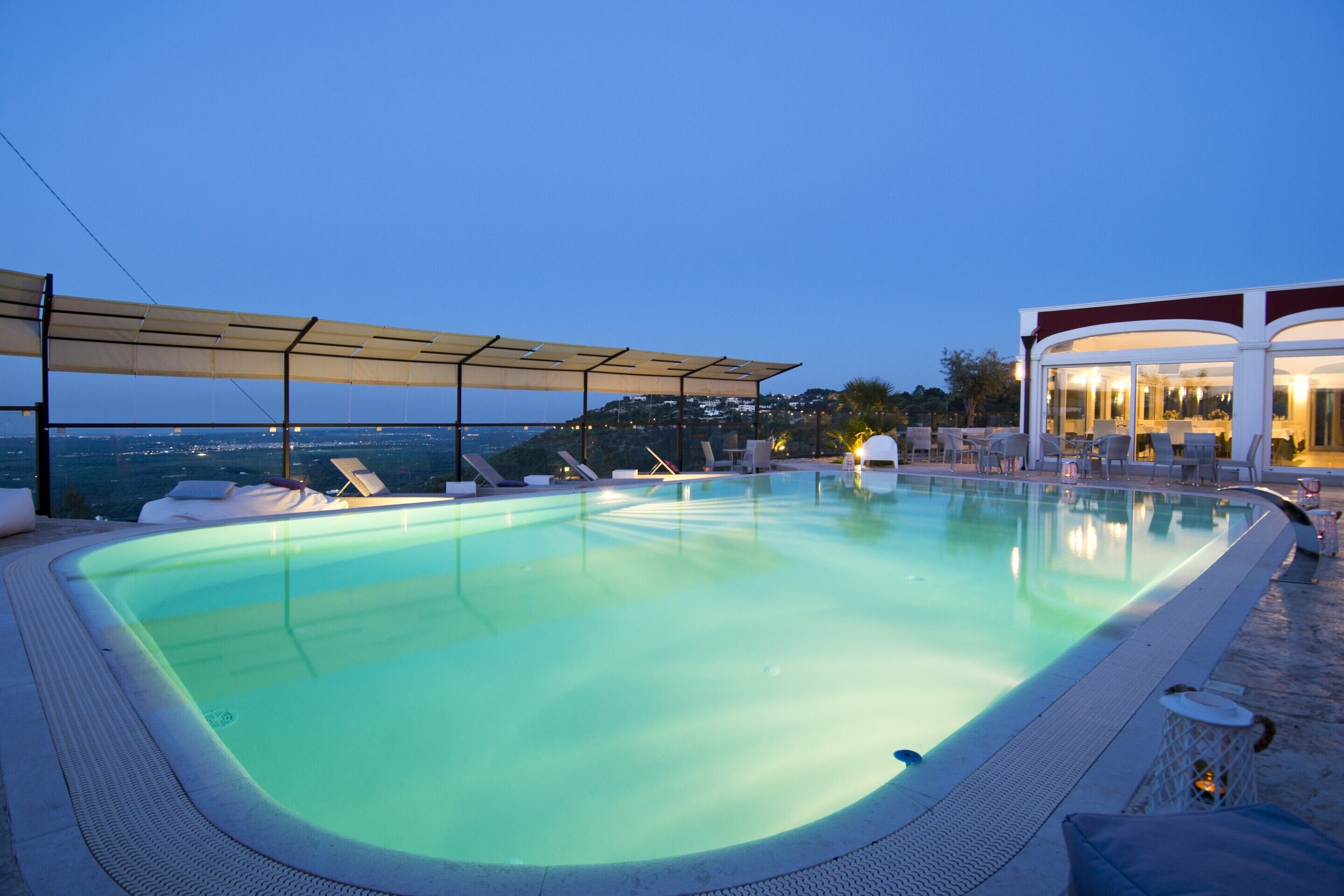 Piscina con sillones de descanso y una zona cubierta, con vistas a un paisaje urbano al atardecer.