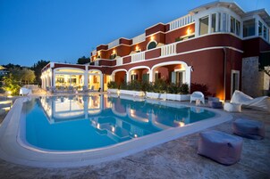 Un hotel grande y moderno con piscina, mobiliario exterior e iluminación destacada en la fachada.