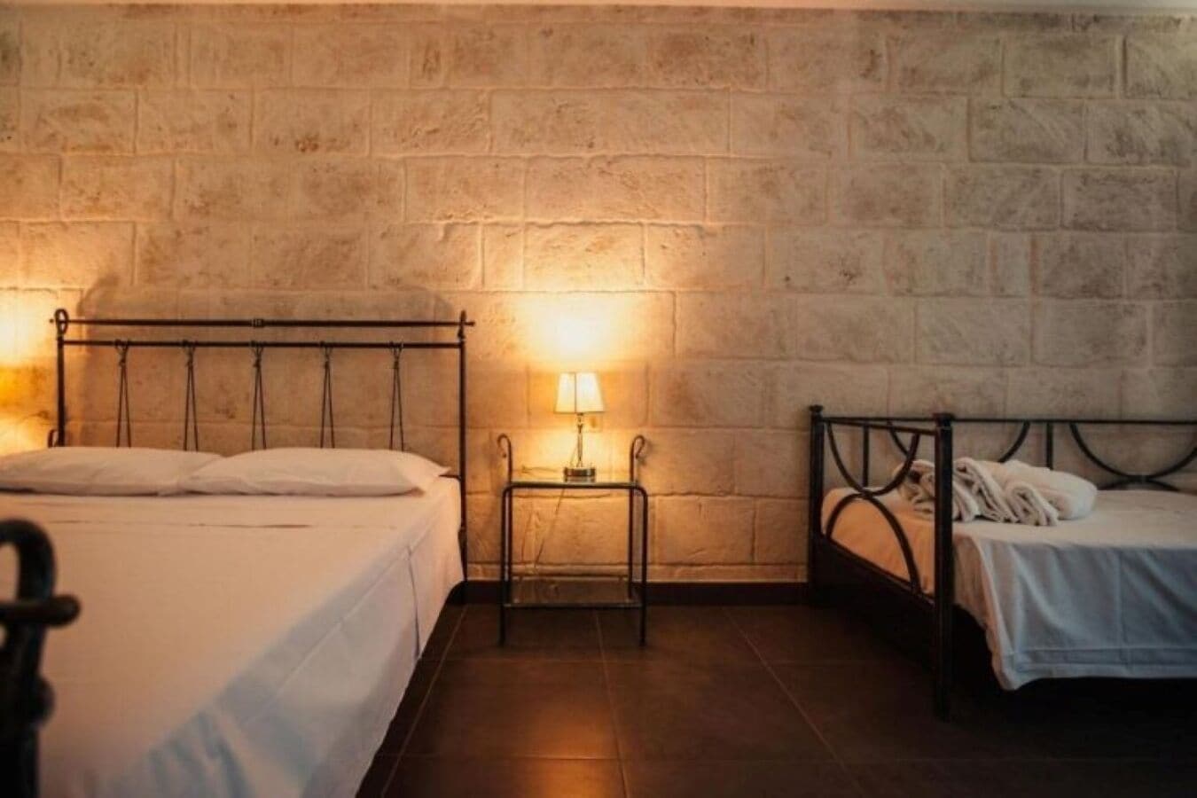 Quarto com duas camas, uma mesinha com abajur e uma parede de pedra.