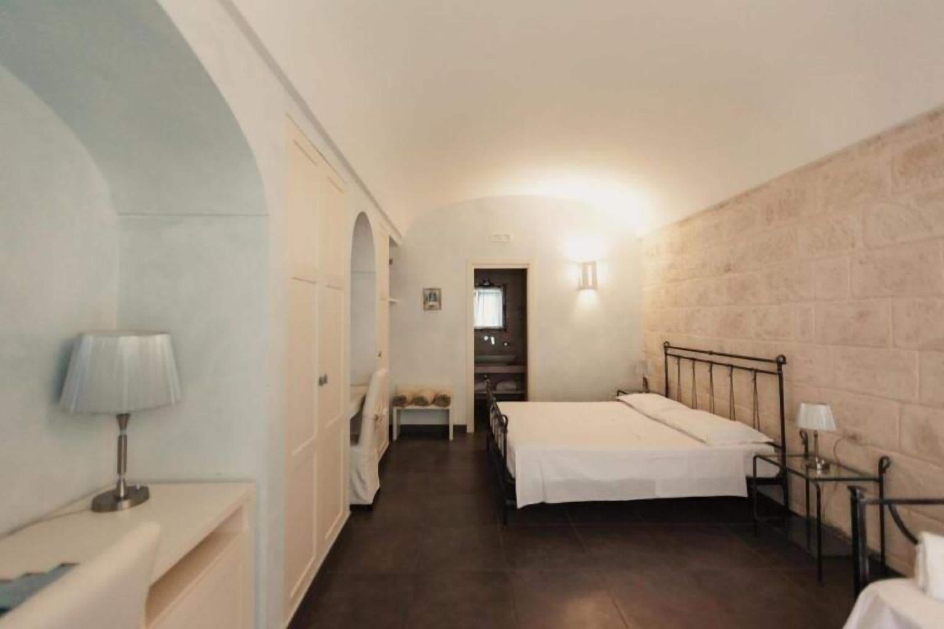 A narrow hotel room with a single bed, a bedside table with a lamp, a small bench, and a view into another room.