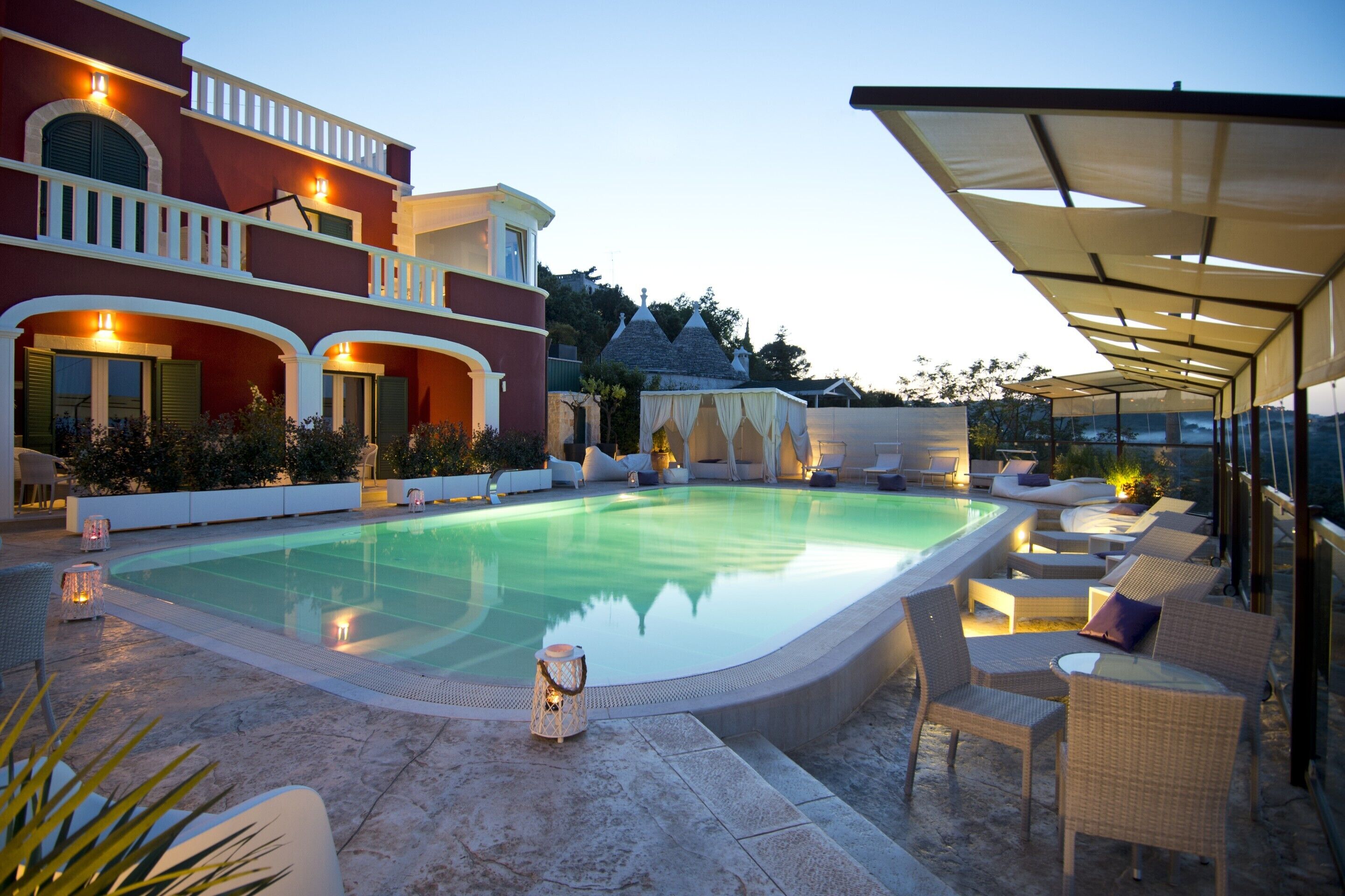 Una piscina con sillones de descanso y sombrillas, rodeada por un edificio con balcones y un patio techado.