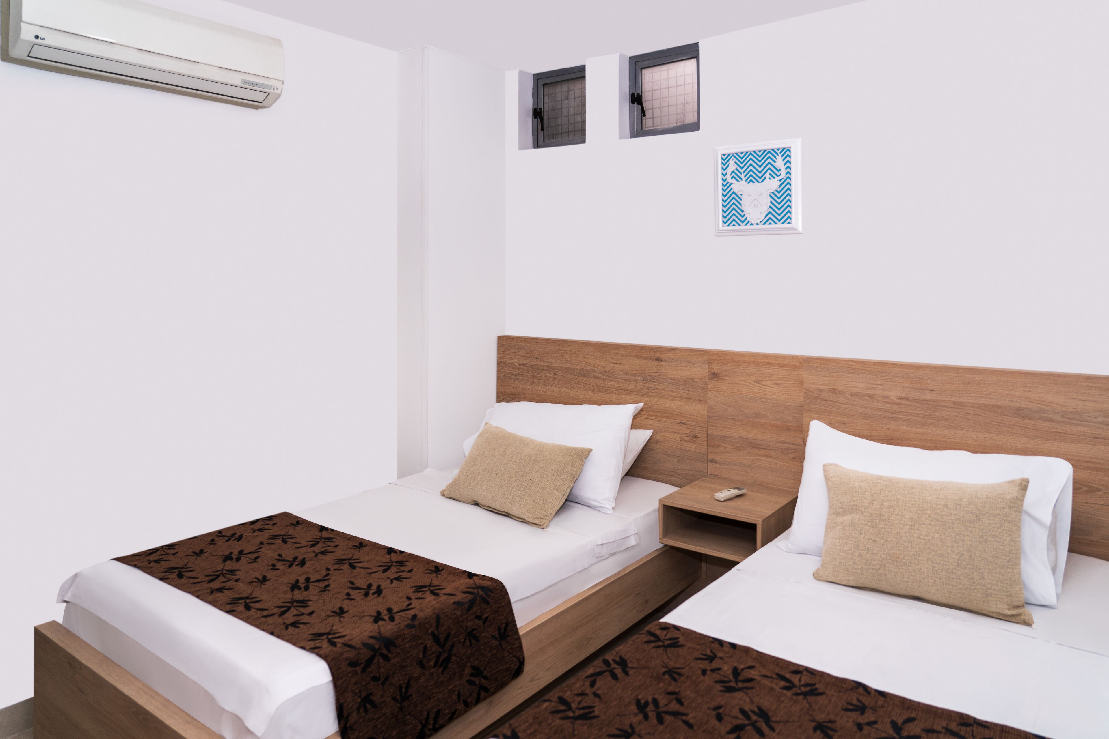 A hotel room with two beds, a wooden headboard, a wall-mounted air conditioner, and two small windows with blinds.
