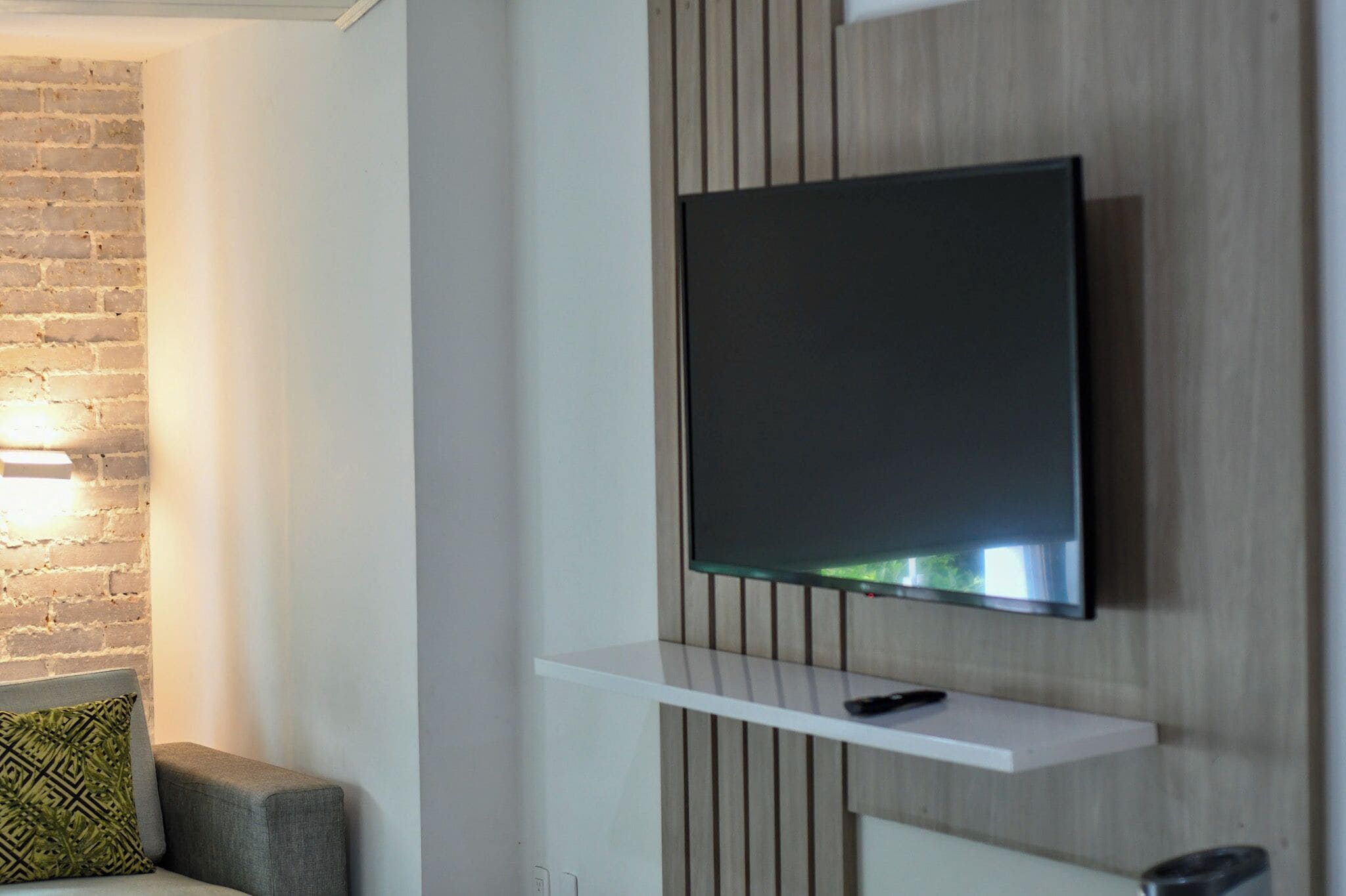Una sala moderna con un televisor de pantalla plana montado en una pared de paneles de madera, un estante blanco debajo y un sofá gris con un cojín estampado.