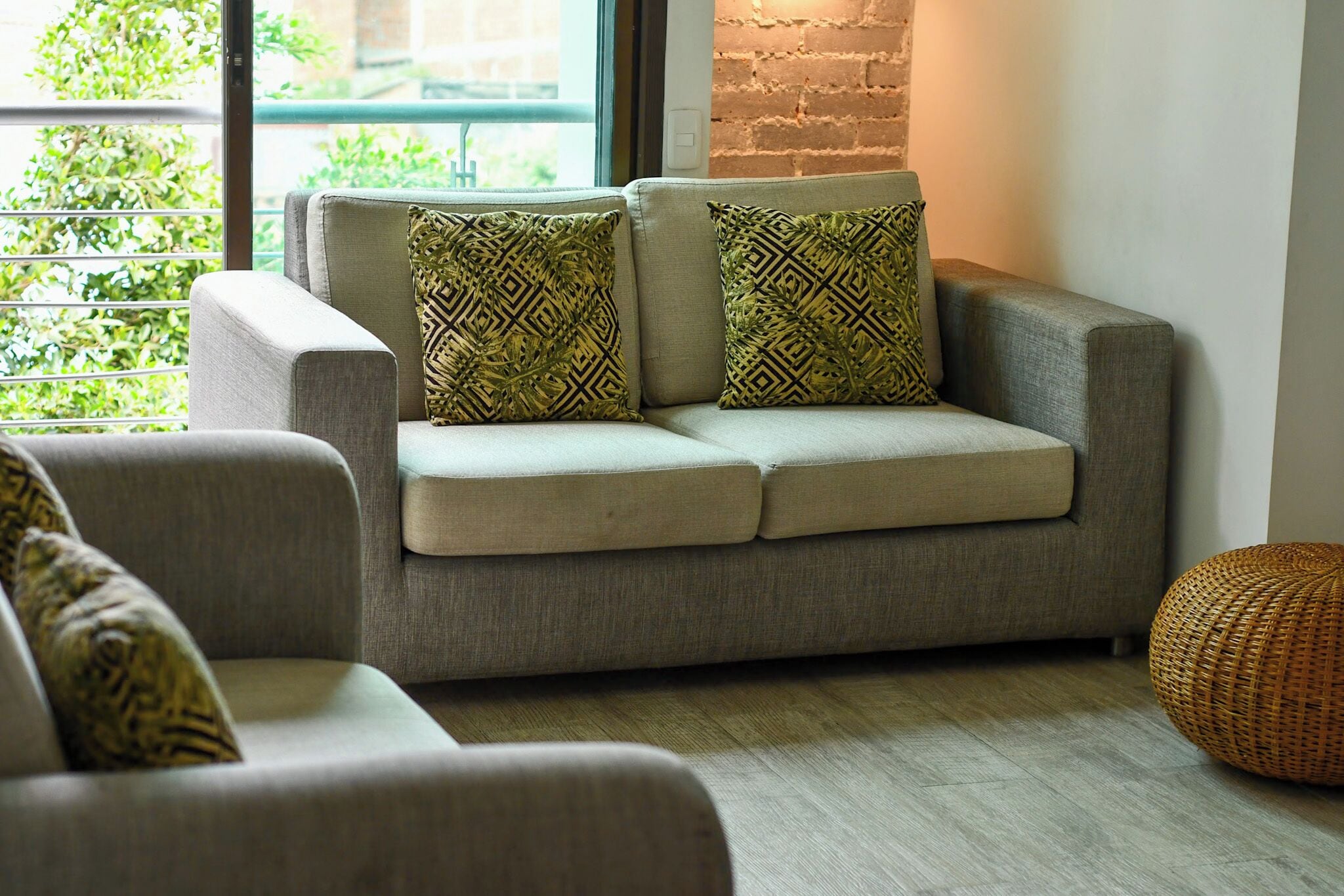 A modern living room with a grey sofa, patterned cushions, a wicker ottoman, and a view through a window.