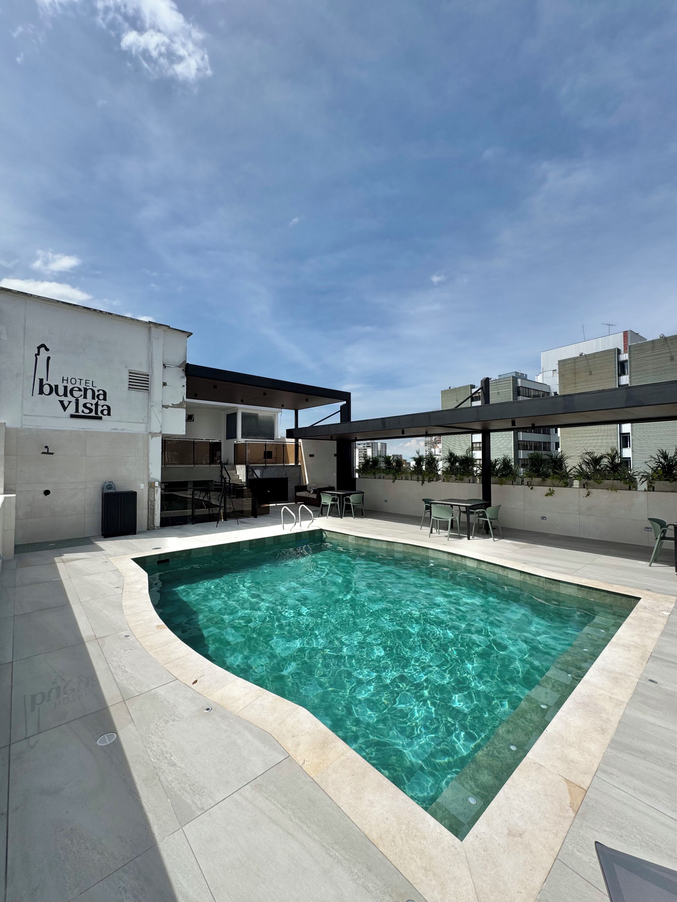 Una piscina con agua cristalina azul, rodeada por un borde de baldosas, y un edificio con un cartel que dice 'HOTEL buena vista'.