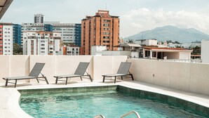 Una piscina en la azotea con sillones, vistas despejadas del paisaje urbano y montañas al fondo.