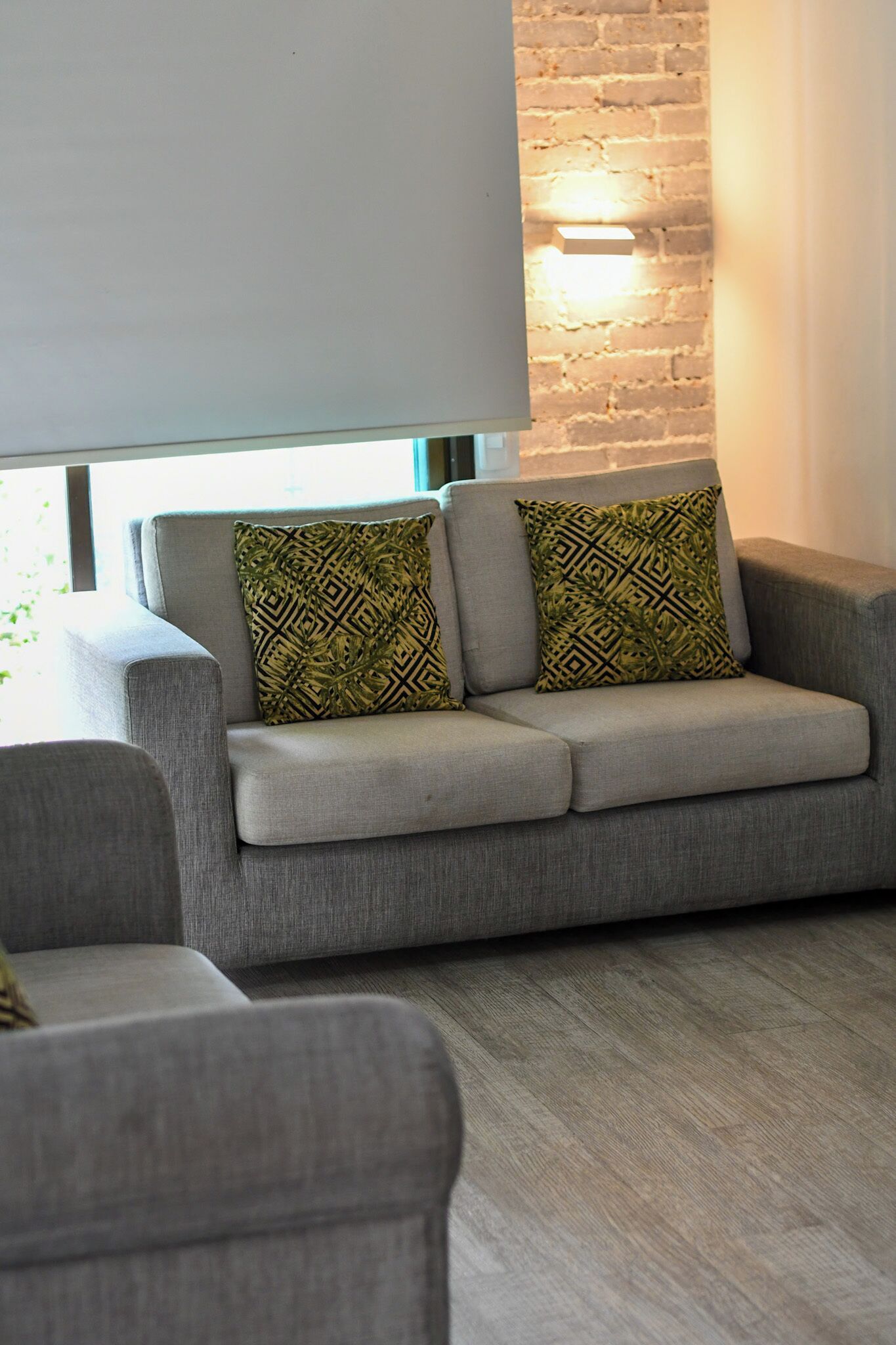 A grey sofa with patterned cushions, a brick wall, and a window with blinds.
