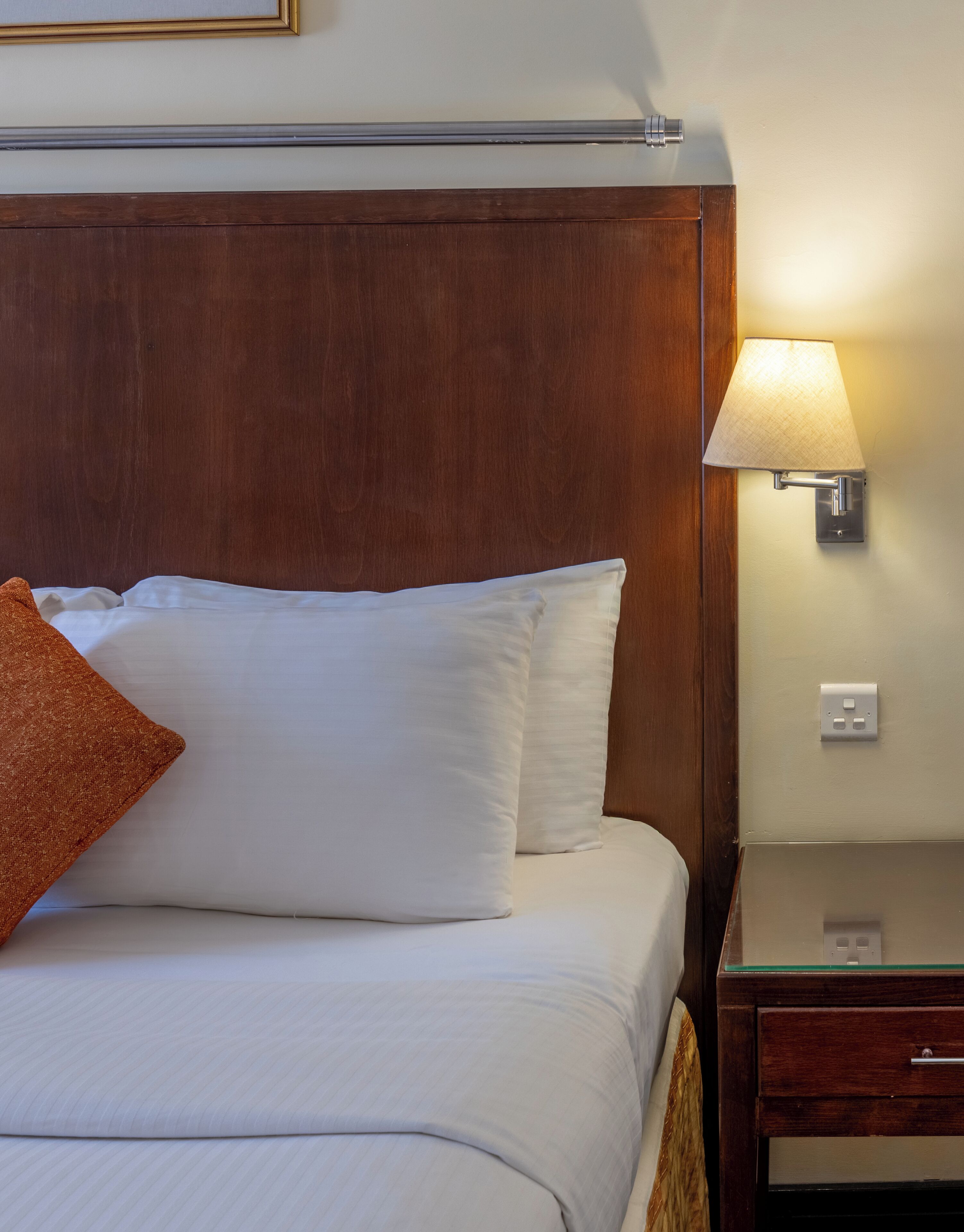 A neatly made bed with white linens and a single brown pillow, a wooden headboard, a bedside table with a lamp, and a wall socket.