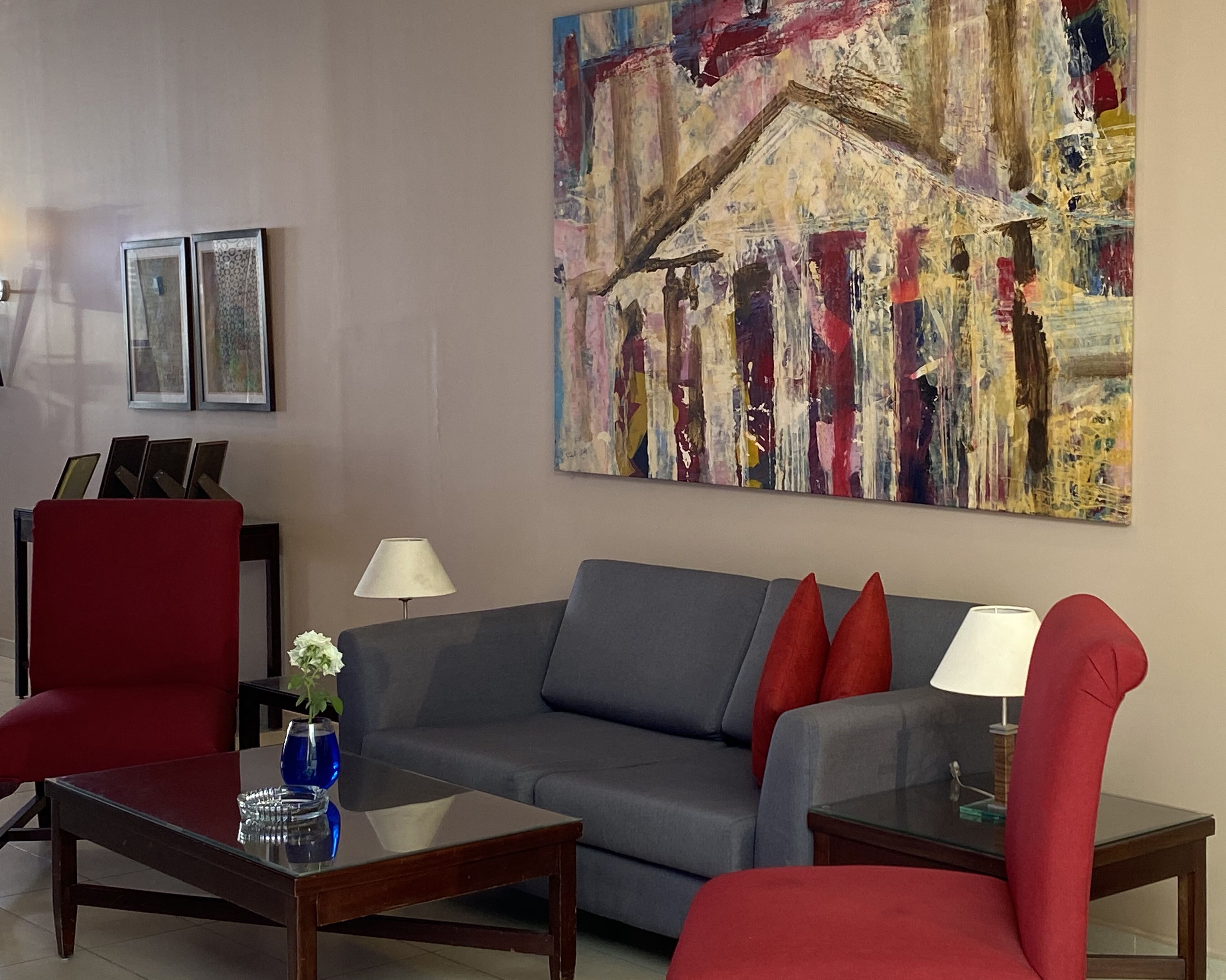 Una sala de estar moderna con un sofá gris, sillones rojos y una mesa de centro. Hay dos lámparas, un jarrón con flores y un cuadro abstracto de gran tamaño en la pared.