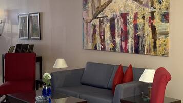 Una sala de estar moderna con un sofá gris, sillones rojos y una mesa de centro. Hay dos lámparas, un jarrón con flores y un cuadro abstracto de gran tamaño en la pared.