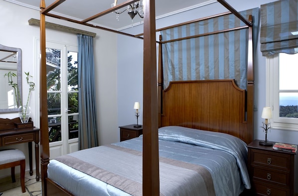 A four-poster bed with a wooden headboard, a bedside table with a lamp, and a window with curtains. - Dafnoudi (Kefalonia)