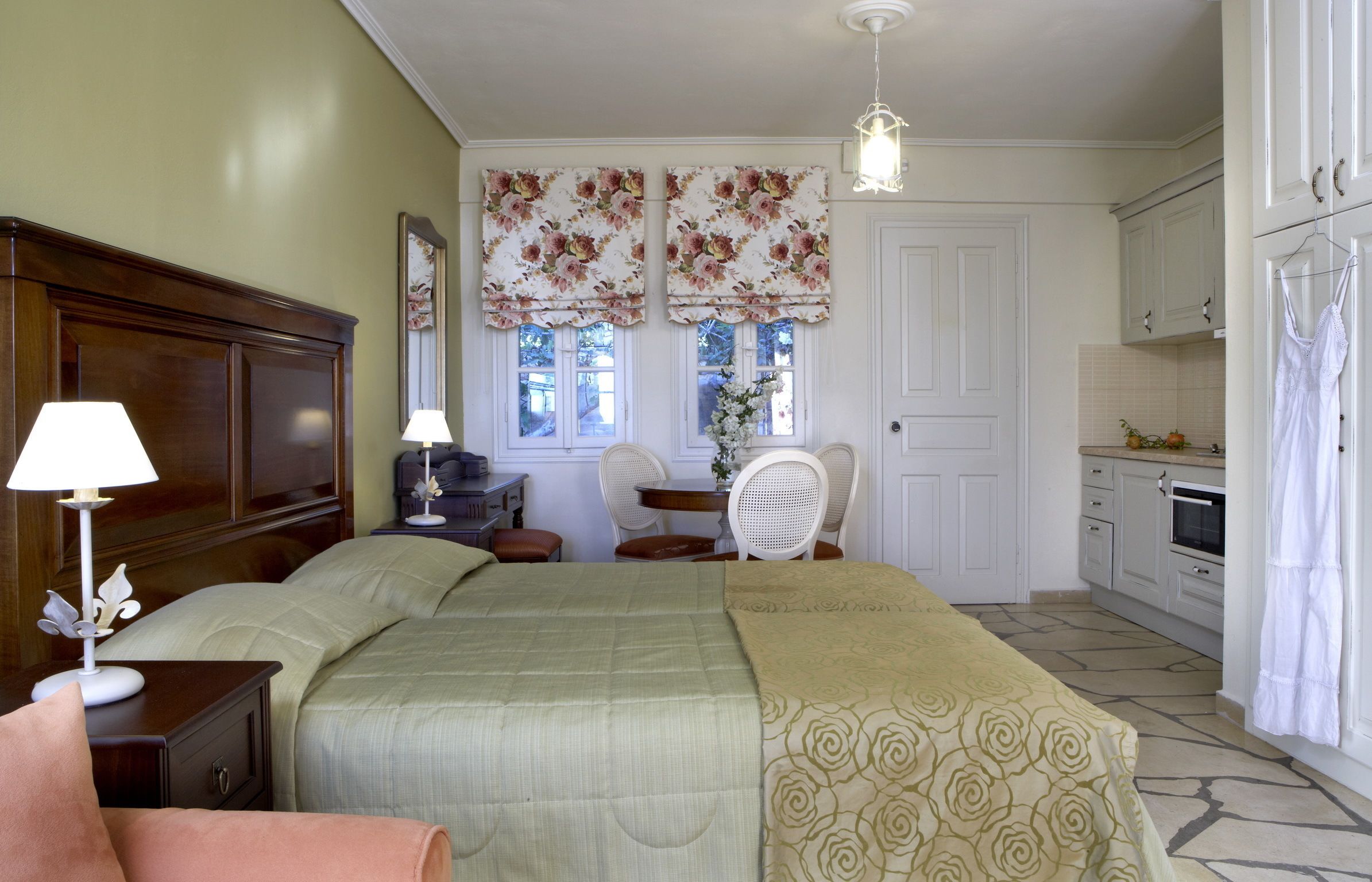 A bedroom with a bed, bedside table, lamp, and a chair. There is a window with floral curtains and a view of the outside.