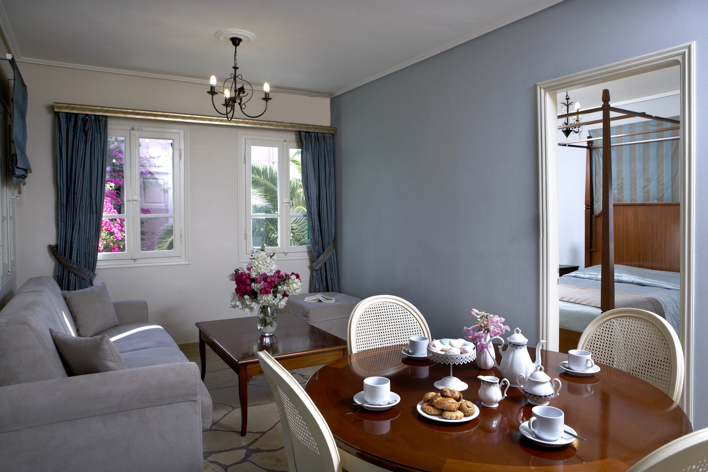 A dining area with a round wooden table set for tea, surrounded by white chairs, a grey sofa, and a window with blue curtains.
