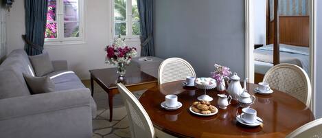 A dining area with a round wooden table set for tea, surrounded by white chairs, a grey sofa, and a window with blue curtains.