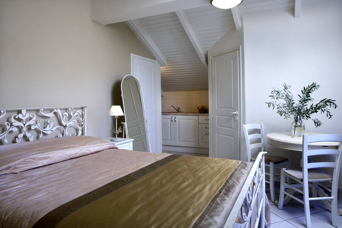 Un dormitorio con cama, mesita de noche, una silla, una pequeña mesa con una planta, y una cocina visible a través de una puerta abierta.