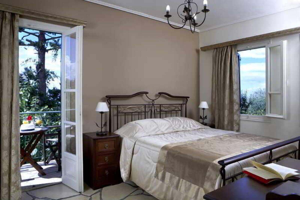 Un dormitorio con cama, lámparas de noche, mesita de noche, ventana con cortinas y balcón con mesa y sillas.