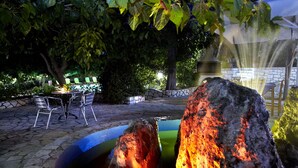 An outdoor patio with stone flooring, metal chairs, and a table. There is a rock feature with a water stream and a fountain. - Dafnoudi (Kefalonia)