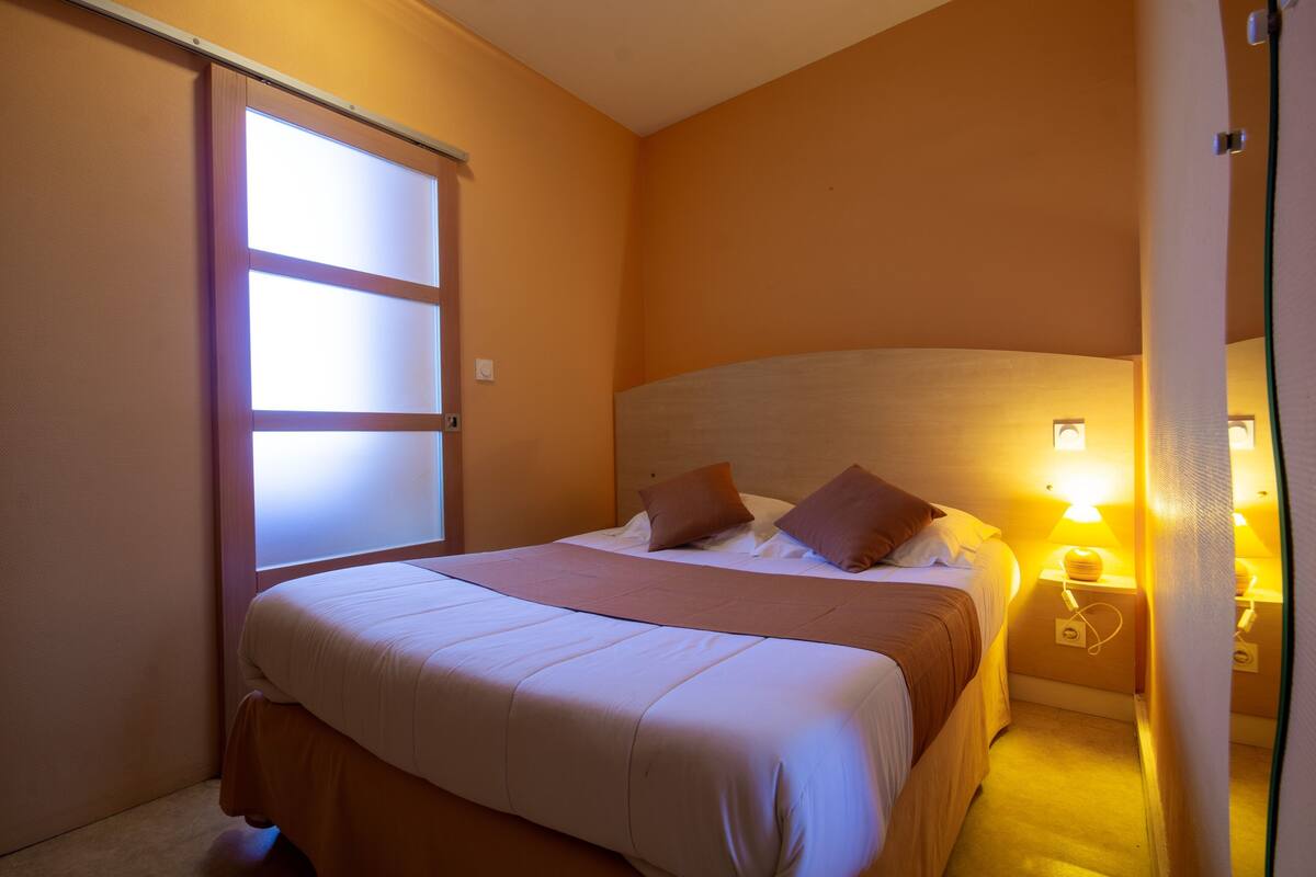Habitación de hotel con cama, mesita de noche con lámpara, ventana con persianas y luz en la pared.