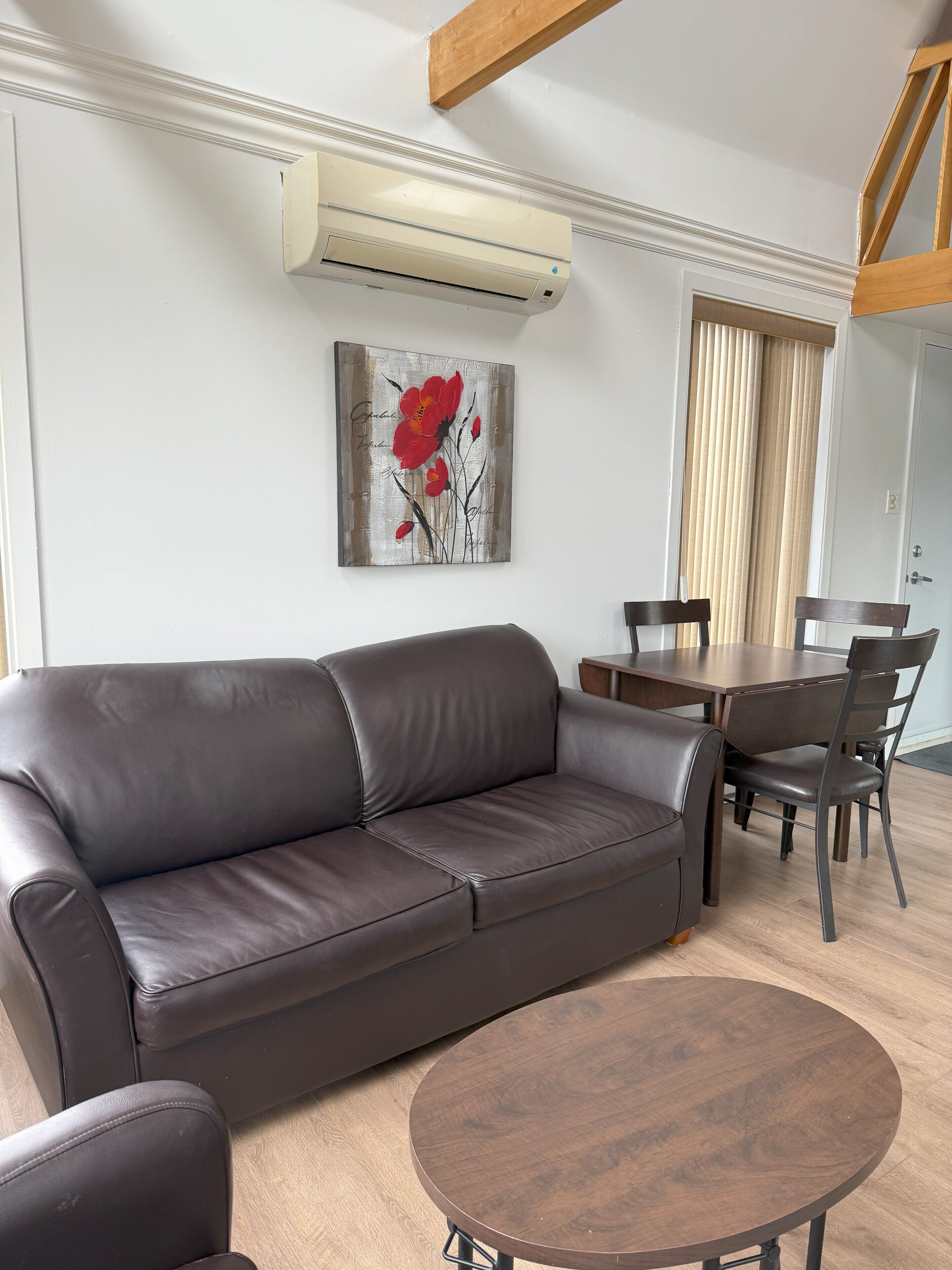 Un salón con un sofá de cuero marrón, una mesa de centro de madera y un comedor con mesa y sillas de madera. Un equipo de aire acondicionado está montado en la pared, y un cuadro de flores rojas cuelga sobre el sofá.