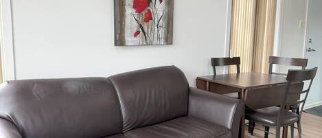 Un salón con un sofá de cuero marrón, una mesa de centro de madera y un comedor con mesa y sillas de madera. Un equipo de aire acondicionado está montado en la pared, y un cuadro de flores rojas cuelga sobre el sofá.