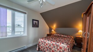 A bedroom with a large window, a ceiling fan, a bed with a quilt, two bedside tables with lamps, and a wooden wardrobe. - Condos Vacances MSA CONDO (Beaupre)