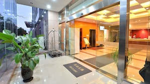 Vestíbulo de hotel con mostrador de recepción, entrada de cristal y una planta en maceta.