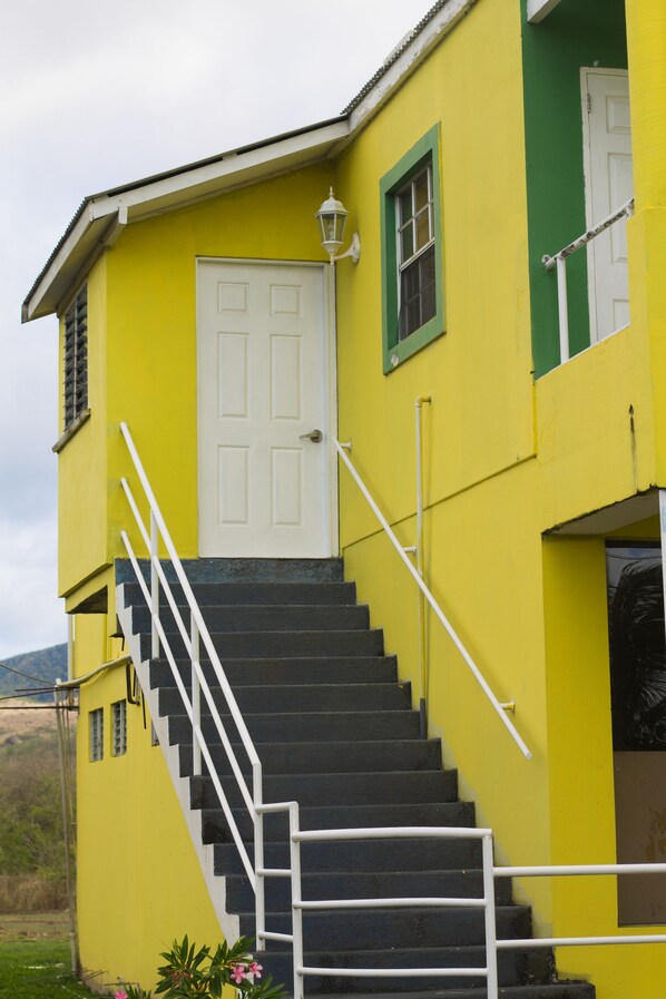 A yellow building with a white door and a staircase with white railings. - Caribbean Holiday Apartments (St. John's)