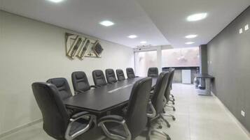 Sala de conferências com uma mesa comprida e várias cadeiras, equipada com iluminação suspensa e unidade de ar condicionado visível.