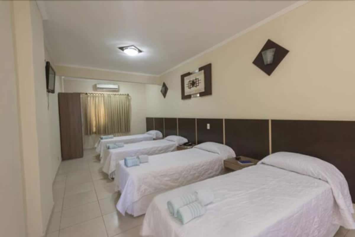 A hotel room with four single beds, a large bed, a headboard, a wall-mounted air conditioner, and a framed picture.