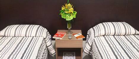 A hotel room with two beds, a nightstand with a vase of flowers, and a small shelf with books.