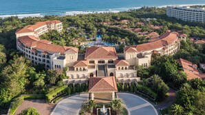 A large resort with a central pool, surrounded by lush greenery and palm trees, with a view of the ocean.