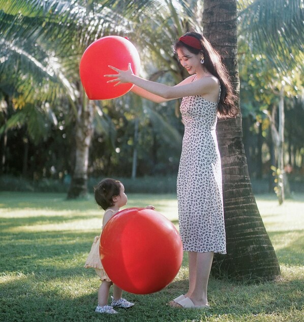Una mujer y un niño jugando con una pelota roja grande en un área verde con palmeras.