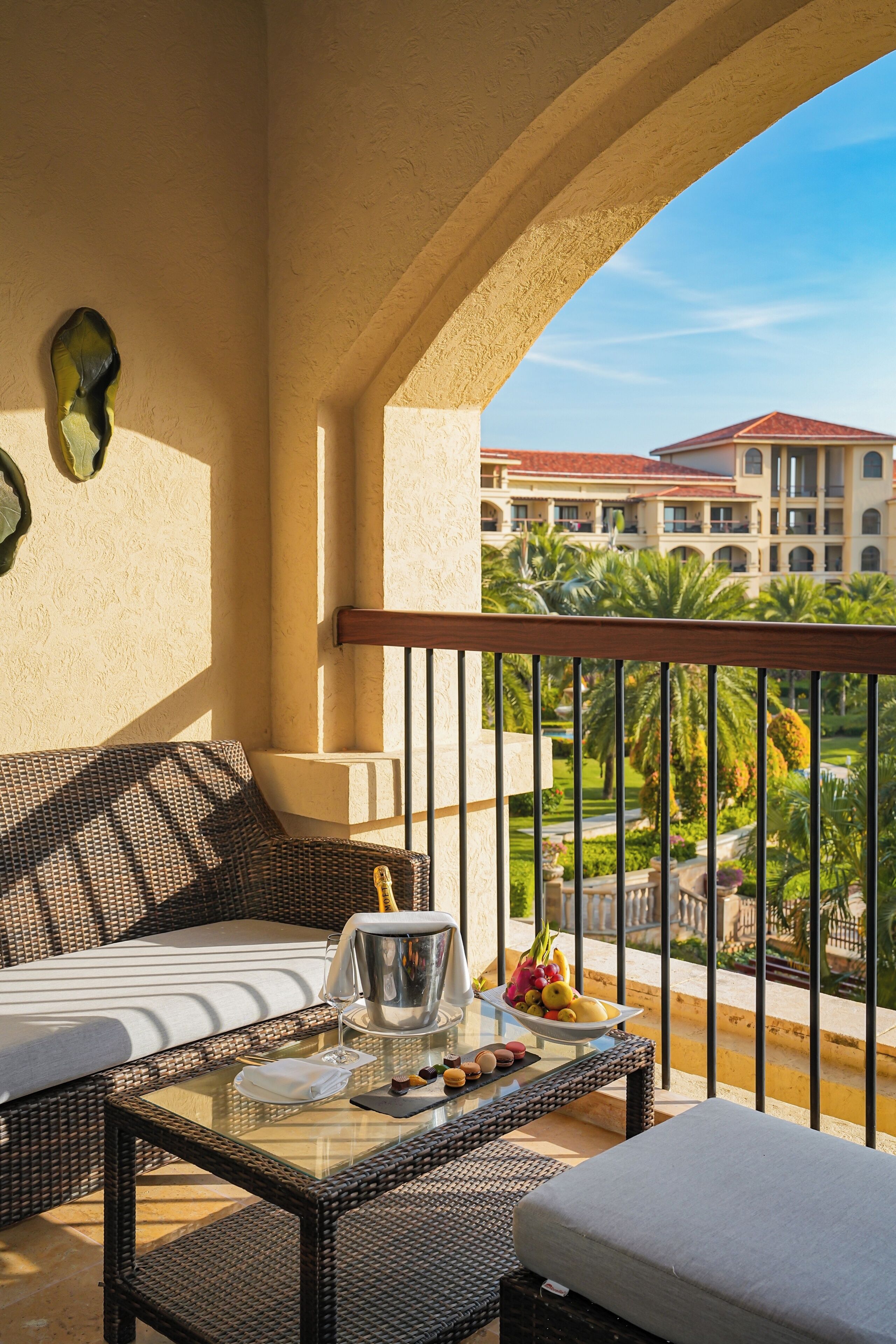 A balcony with wicker furniture, a table with a pitcher and fruit, and a view of a building with a red roof.