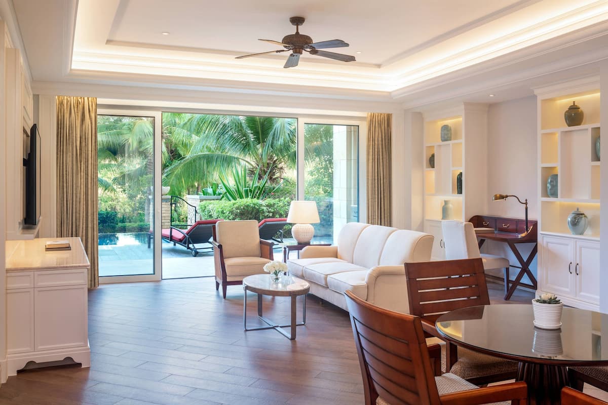 A spacious living area with a sofa, chairs, a coffee table, and a dining table. There is a ceiling fan, a large window with a view of greenery, and a cabinet with decorative items.
