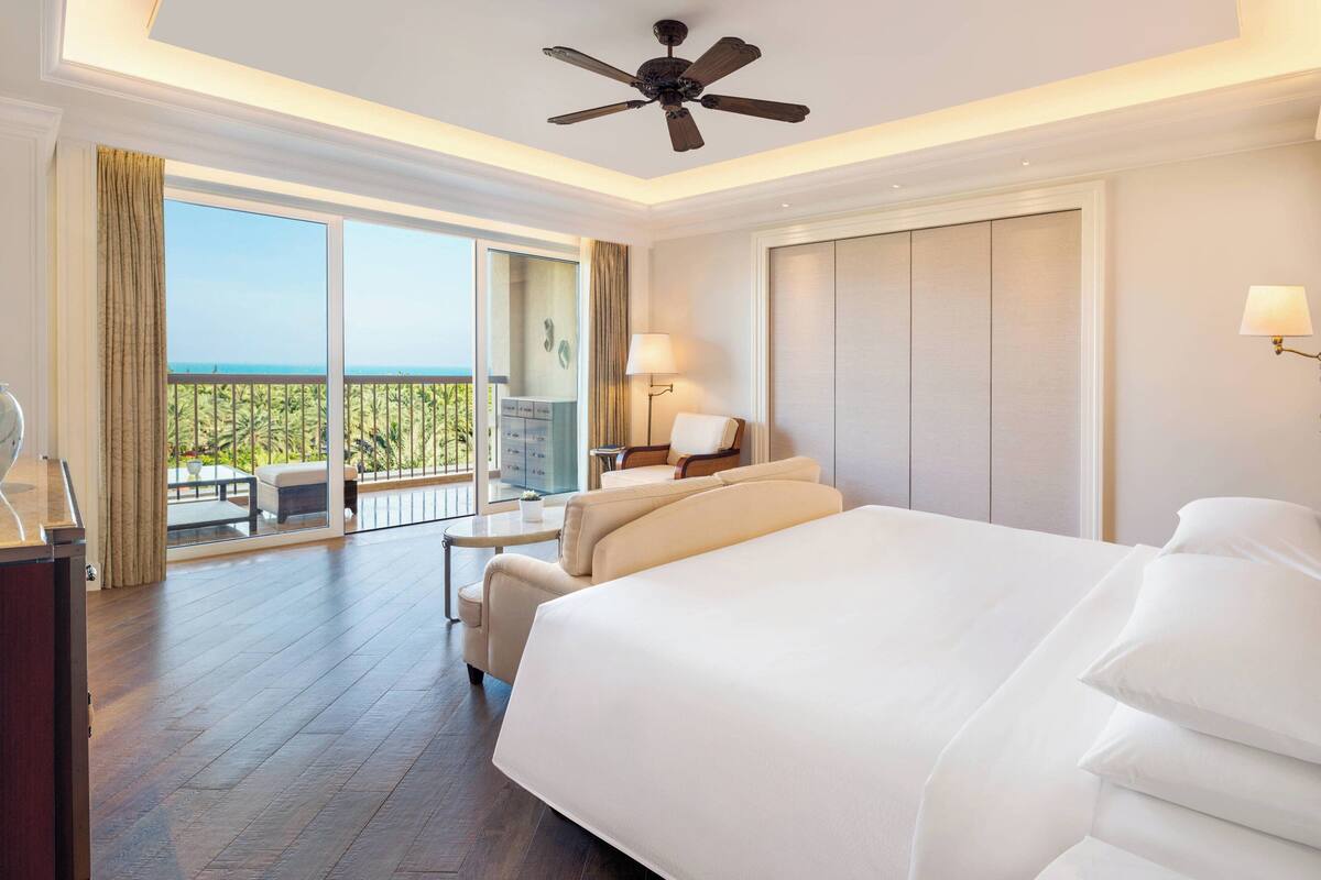 A hotel room with a large bed, a ceiling fan, a balcony with a view, and a desk with a lamp.
