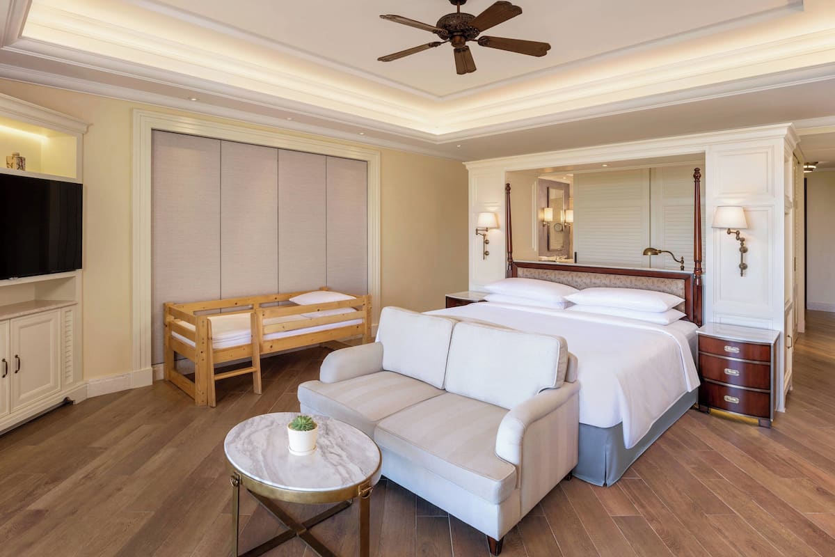 A hotel room with a bed, a sofa, a TV, a wooden bench, a small table with a plant, and a ceiling fan.