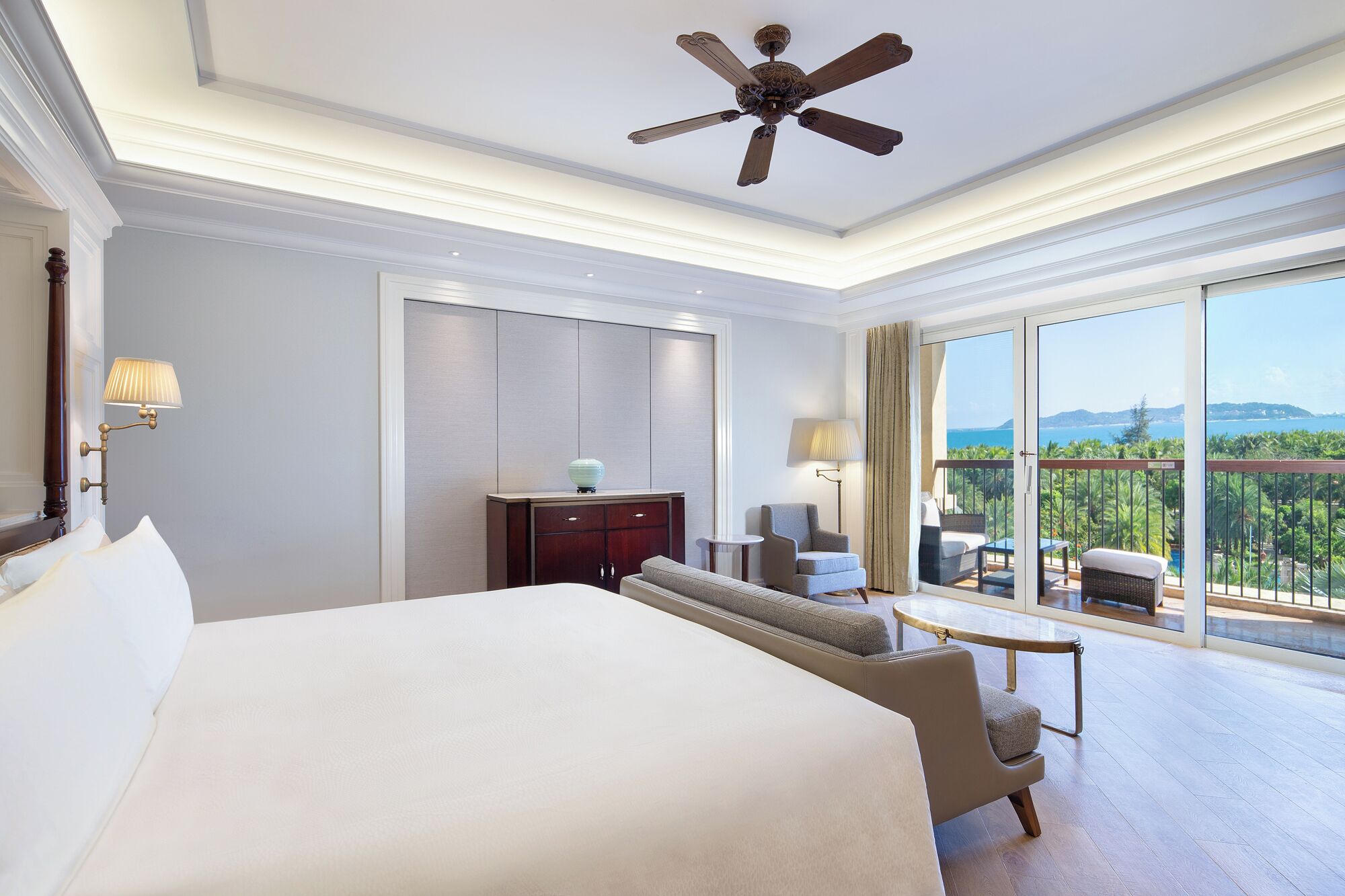 A hotel room with a large bed, a desk, a chair, a balcony with a view, and a ceiling fan.