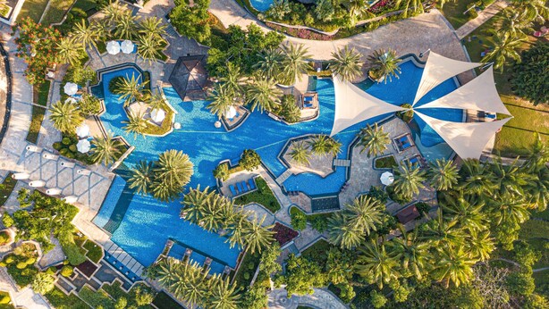 An aerial view of a resort with multiple swimming pools, palm trees, and sun umbrellas.